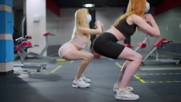 Zijaanzicht van twee fitte sportvrouwen in Covid gezichtsmaskers die in de sportschool squats doen. Gemotiveerde slanke jonge blanke vrouwen die binnen trainen op een coronavirus pandemie. Sport en nieuw normaal. — Stockvideo