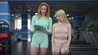 Spor salonunda yürüyen ve tablet üzerinde sosyal medyada sörf yapan kendine güvenen iki genç kadının ön görüntüsü. Güzel sarışın ve kızıl saçlı beyaz tenli kadınların portresi. Eğitimden önce geziniyorlar..