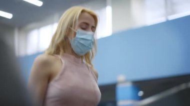Beyaz kadın portresi, Covid-19 yüz maskesi takmış boyalı sarı saçlı Coronavirus salgınında koşu bandında koşan. Zayıf, formda bir sporcu kadın içeride koşuyor. Kardiyo eğitimi.