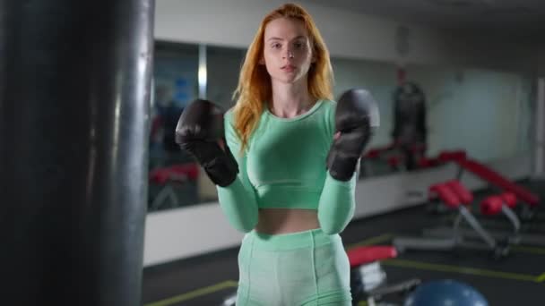 Середній постріл сильної жінки, яка б'є боксерські рукавички, виставляючи їх у спортзалі в приміщенні. Портрет рудої кавказької тоненької спортсменки, яка дивиться на камеру з впевненим виразом обличчя. Жіноча концепція боксу. — стокове відео