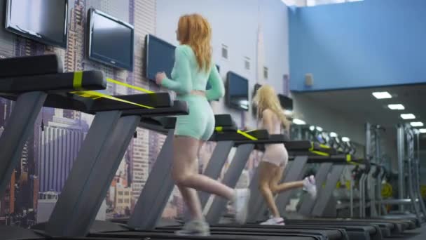 코로나 바이러스가 유행하는 헬스 장에서 러닝 머신 위에서 달리고 있는 두 명의 건강 한 스포츠 여성의 넓은 샷. 빨간 머리와 금발 백인 여성들 이 실내에서 조깅을 하고 있습니다. 사회적 거리와 사업의 재개장. — 비디오