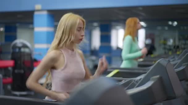 Medium-shot portrett av en ung, vakker hvit kvinne som jogger på tredemøllen som en utydelig, rødhåret sportskvinne som løper i bakgrunnen. Selvsikre kvinner som jobber innendørs. – stockvideo