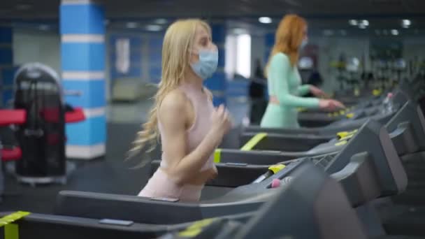 Πλάγια άποψη πορτρέτο του ξανθιά νεαρή ταιριάζει γυναίκα σε coronavirus μάσκα προσώπου τρέχει στο διάδρομο ως θολή κοκκινομάλλα αθλητικογράφος άσκηση τζόκινγκ στο παρασκήνιο. Σλιμ γυναίκες κατάρτιση σε Covid-19 πανδημία. — Αρχείο Βίντεο