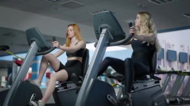 Sarışın, zinde bir kadın, kızıl saçlı arkadaşıyla egzersiz bisikletleri üzerinde konuşuyor. Spor salonunda spor yapan zayıf, güzel beyaz kadın portresi. Sağlıklı yaşam tarzı ve kadınlık.