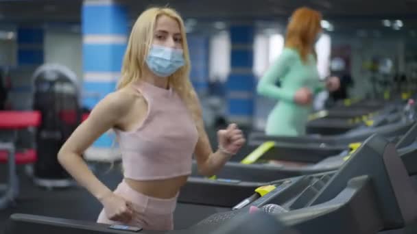 Coronavirus yüz maskesi takmış neşeli, kendine güvenen sporcu kadın kameraya bakıyor ve spor salonunda koşu bandında koşuyor, arka planda koşan kızıl saçlı kadın.. — Stok video