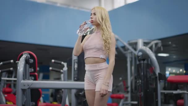 Cansada joven atleta caucásica en forma bebiendo agua refrescante de pie en el gimnasio y girando a la cámara. Retrato de una mujer guapa rubia en ropa deportiva rosa posando después del entrenamiento en interiores. — Vídeo de stock