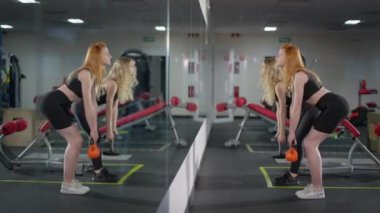Spor salonundaki ağırlık kaldıran kendine güvenen sporcu kadınların yan görünüşü aynaya yansıyor. Pozitif gülümseyen kızıl saçlı ve bin yıllık sarışın kapalı alanda çalışan beyaz kadınlar. Ağırlık kaldırmak ve spor.