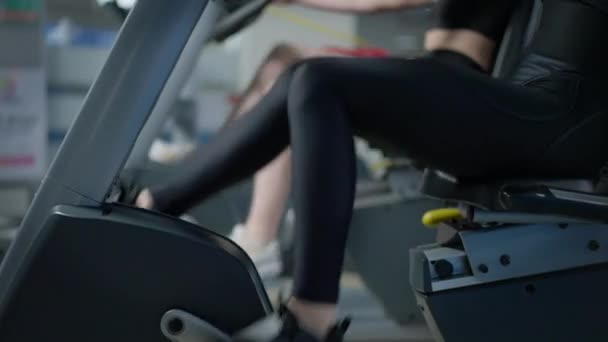 Side view benen van slanke vrouwen rijden stationaire fiets in de sportschool. Twee onherkenbare blanke fitte sporters trainen binnen op een trainingsmachine. Sportief leefstijlconcept. — Stockvideo