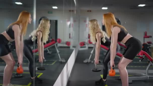 Liten, rødhåret sportskvinne som snakker med blond venn i gymmen og ser speilvendt på refleksjoner. Portrett av fine hvite kvinner som trener innendørs. Vektløfting og idrettskonsept – stockvideo