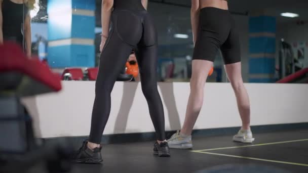 Achteraanzicht van twee slanke, prachtige sporters die hurken met gewichten in de sportschool. Fit blanke vrouwen heffen gewichten training binnen in de voorkant van de spiegel. Workout en bodybuilding. — Stockvideo