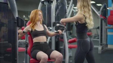 Spor salonundaki pazılı eğitim makinesini müşteri için kullanan antrenör. Kızıl saçlı zayıf kadın ekipmanların içinde oturmuş antrenman yapıyor ve içeride müşterileri destekliyor. Spor ve hizmet..