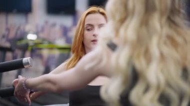 Yakın plan kızıl saçlı sporcu kadın kişisel antrenörüyle el kaslarını çalıştırıyor ve arka plandan geçen dövmeli adama yardım ediyor. Spor salonunda egzersiz yapan Kafkas kökenli bir kadının portresi..