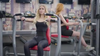 Sarışın, zayıf kadının geniş açılı portresi. Biseps kaslarını ekipman üzerinde çalıştırıyor. Arka planda egzersiz makinesinde çalışan kızıl saçlı sporcu kadın..