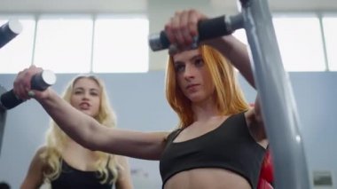 Özenle hazırlanmış kızıl saçlı sporcu kadın arka planda neşeli, bulanık kişisel antrenörü olan ekipman üzerinde el kasları çalıştırıyor. Sıska bir kadın spor salonunda çalışıyor..