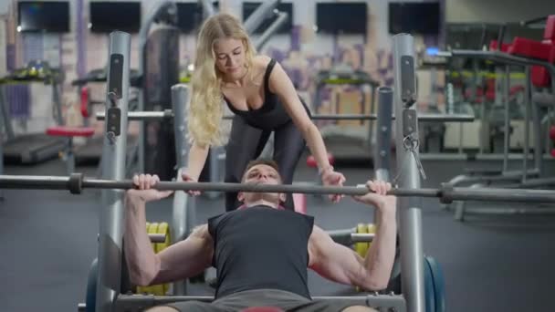 Entrenador personal delgado y rubio que ayuda al deportista a levantar pesas en el gimnasio acostado en el banco de ejercicios. Retrato de una joven caucásica sonriente respaldando a un hombre fuerte trabajando en interiores. — Vídeos de Stock