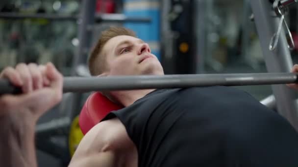 Primer plano de deportista guapo motivado levantando la barra exhalando tumbado en el banco de ejercicio en el gimnasio. Confiado concentrado caucásico joven entrenar en el interior. — Vídeo de stock