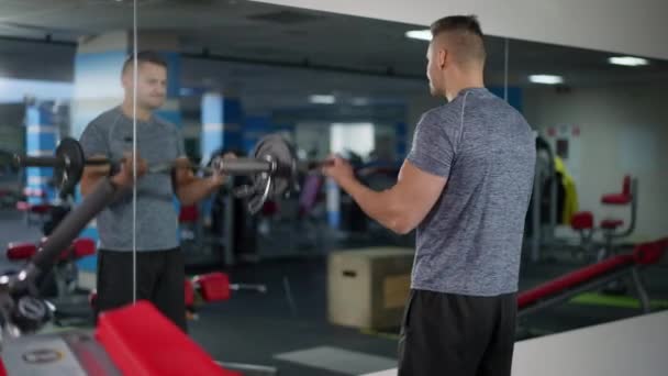 Bakspeilet på en sterk idrettsmann som løfter tunge bjeller og reflekterer i speilet i gymsalen. Selvsikker, motivert hvit mann som trener innendørs. Vektløfting og livsløpskonsept. – stockvideo