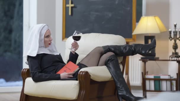 Широкий снимок расслабленной женщины в костюме монахини и высоких кожаных сапогах, сидящей на удобном кресле со Священной Библией и смотрящей на бокал вина. Боковой вид беззаботной провокационной дамы в помещении. — стоковое видео