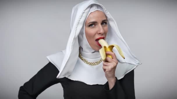 Lákavá běloška v kostýmu jeptišky, která jí banán a usmívá se na kameru. Portrét vášnivé svůdné dámy s červenou rtěnkou těší lahodné ovoce pózování na šedém pozadí. — Stock video