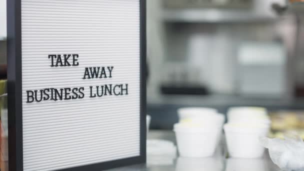 Close-up Annuncio pranzo di lavoro take away con imballaggio sfocato cuoco contenitori usa e getta con il cibo in background. Donna irriconoscibile in guanti che prepara ordini in mensa all'interno. — Video Stock