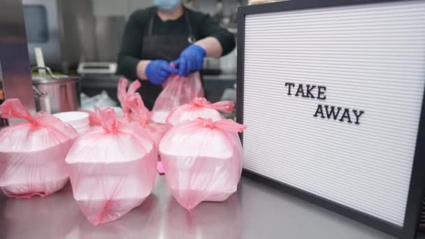 Close-up Llevar mensaje con alimentos empacados en el mostrador y borrosa voluntaria que trabaja en segundo plano. Mujer tipo caucásica preparando bocadillos para personas sin hogar en la cocina comercial. — Vídeos de Stock