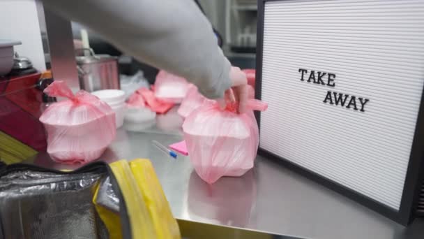 Mensajero irreconocible poner embalado llevar recipientes de comida en la bolsa con el cocinero trabajando en segundo plano. Joven hombre caucásico trabajando entregando el almuerzo. Concepto de trabajo y cena. — Vídeos de Stock