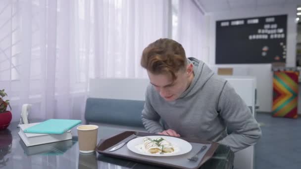 Estudiante masculino oliendo delicioso postre en cantina sonriente. Retrato de un joven guapo caucásico sentado en la cafetería con almuerzo. Estilo de vida y concepto de ruptura. — Vídeos de Stock