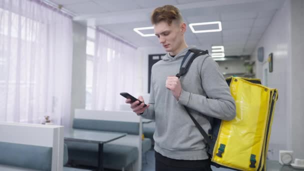 Συγκεντρωμένος νεαρός άνδρας στέκεται στο καφέ με κίτρινη τσάντα παράδοσης τροφίμων έλεγχο online παραγγελίες στο smartphone, και κοιτάζοντας την κάμερα. Πορτρέτο ενός όμορφου καυκάσιου μαθητή σε εσωτερικούς χώρους. — Αρχείο Βίντεο