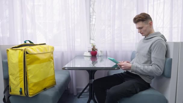 Boční pohled portrét veselého mladého kavkazského kurýra kontrolujícího online objednávku v tabletu při pohledu na usmívající se kameru. Žlutý tepelně izolovaný batoh stojící na gauči v jídelně. — Stock video