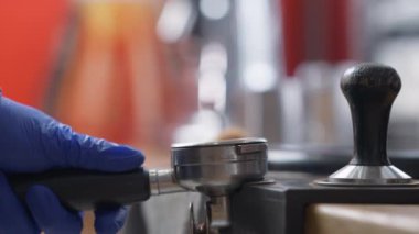 Barista, grup tutamağında kahve tozu bastırırken yakın plan sıkıştırıyor. Tanımlanamayan beyaz eldivenli adam kafeteryada sıcak içecek hazırlıyor..