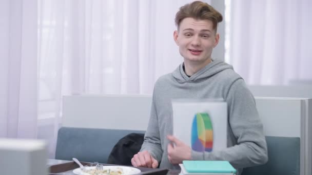 Inteligente milenario joven estudiante varón posando con diagrama en la cantimplora sentado a la mesa con comida. Retrato del hombre caucásico sonriente positivo mirando la cámara en la hora del almuerzo en la cafetería. — Vídeos de Stock