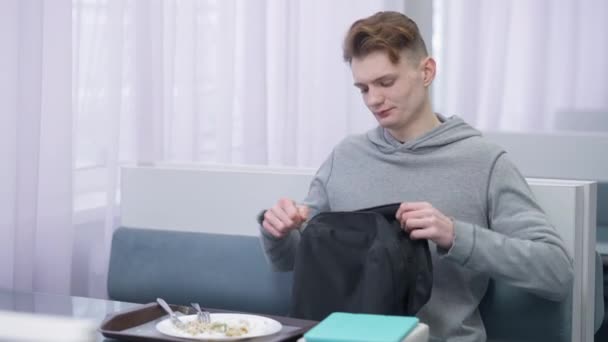 Joven estudiante inteligente guapo empacar mochila y salir de la cafetería. Retrato de un hombre caucásico inteligente concentrado poniendo libros y tabletas en una bolsa alejándose de la cantina. Concepto de estilo de vida. — Vídeos de Stock