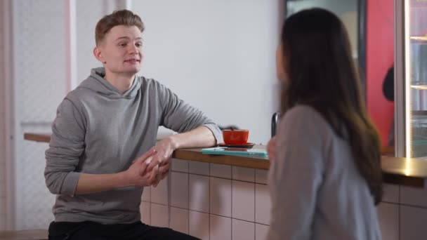 Ung positiv kaukasisk pojkvän talar med flickvän i elev matsal sitter vid disken. Porträtt av stilig självsäker man leende chatta med kvinna inomhus i cafeterian. Dejtingkoncept. — Stockvideo