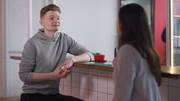 Joven novio positivo caucásico hablando con su novia en la cantina de estudiantes sentado en el mostrador. Retrato de un hombre guapo y confiado sonriendo charlando con una mujer en el interior de la cafetería. Concepto de citas. — Vídeo de stock