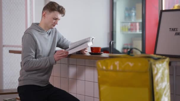 Chytré vstřebané kavkazské tisícileté studentské čtení knihy sedí u jídelního pultu s rozmazaným žlutým sáčkem na jídlo na přední straně. Inteligentní mladý muž učení v práci přestávka uvnitř. — Stock video