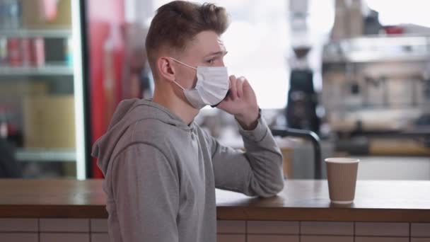 Vista lateral de hombre joven en coronavirus mascarilla hablando por teléfono de pie en el mostrador de la cafetería con taza de café de papel desechable. Retrato de estudiante caucásico en cafetería en pandemia de Covid-19. — Vídeos de Stock