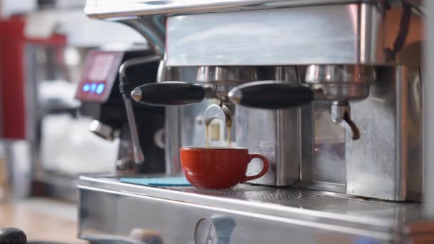 Café espresso recién hecho vertiendo en la taza de café rojo de la máquina de café. Proceso de elaboración de café en cafetería o restaurante en interiores. Barista preparando sabrosa bebida caliente aromática por la mañana en la cafetería. — Vídeo de stock