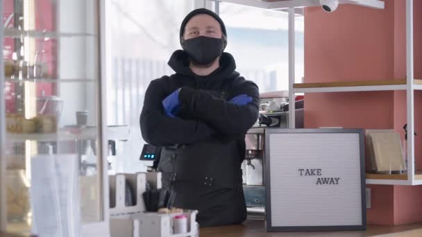 Joven caucásico de confianza en Covid mascarilla cruzando las manos mirando la cámara de pie en el mostrador de la cafetería con el anuncio de Take away. Concepto de reapertura de la industria alimentaria y de bebidas ante la pandemia. — Vídeo de stock