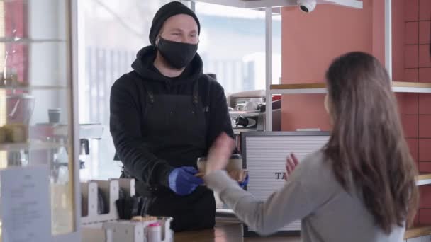 Bartender in coronavirus face mask passing order to client leaving cafe. Portrait of Caucasian young man serving buyer in cafeteria with take away coffee on lockdown re-opening and looking at camera. — Stock Video