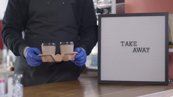 El hombre irreconocible en los guantes que estiran llevan las tazas de café a la cámara. Varón barista caucásico mostrando bebida caliente en taza de papel desechable en el interior de la cafetería. — Vídeo de stock