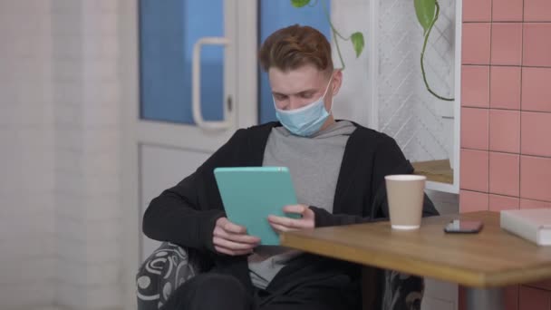 Portrait de jeune homme positif dans le masque facial Covid surfer sur les médias sociaux sur tablette souriant. Étudiant homme millénariste caucasien assis à table dans un café avec tasse de café profiter de loisirs sur le coronavirus. — Video