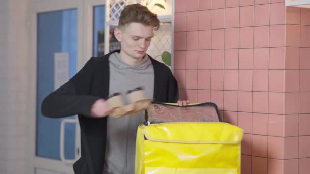 Confiado joven mensajero poniendo llevar café en la mochila de entrega de comida con cremallera bolsa amarilla y mirando a la cámara. Retrato de hombre caucásico guapo entregando bebida caliente de café posando en el interior. — Vídeos de Stock