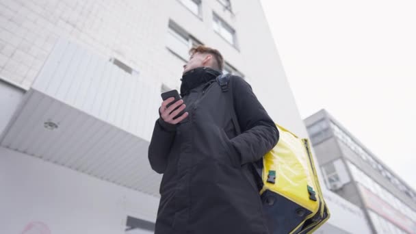 Вид снизу на ориентированного молодого курьера, стоящего на улице с желтой сумкой для доставки еды, проверяющего адрес в приложении для смартфонов. Портрет концентрированного кавказца, ищущего клиента. — стоковое видео