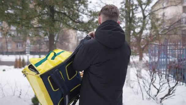 Visszapillantás a fiatalemberre, amint felakasztja az ételszállító hátizsákot, és elsétál a téli napon a szabadban. Magabiztos kaukázusi férfi futár sárga termáltáskával ételt és italt szállít városi városban. — Stock videók