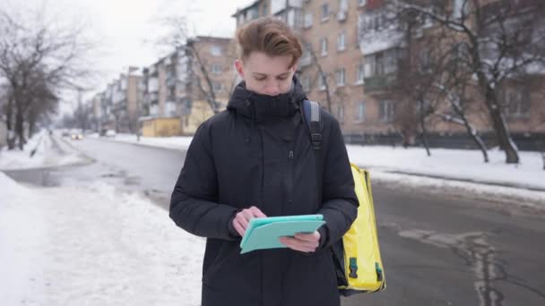 Masculin student mesagerie on-line tabletă de închidere și de mers pe jos cu sacul de livrare alimente galbene. Portretul unui bărbat caucazian concentrat care lucrează în aer liber în timpul iernii . — Videoclip de stoc