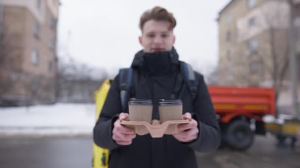 Blurred young courier stretching disposable paper coffee cups at camera standing outdoors on cold winter day. Caucasian man delivering hot drink. Clients point of view. — Stock Video