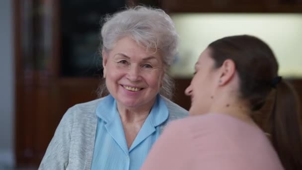 Porträt einer glücklichen Seniorin, die lächelnd in die Kamera blickt, während die junge Enkelin ihre Großeltern umarmt. Positive kaukasische Rentnerin posiert zu Hause mit Enkelkind. Konzept zur Familienzusammenführung. — Stockvideo