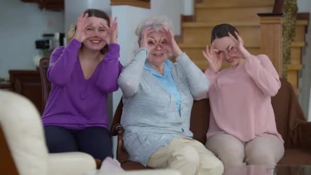 Mulheres felizes alegres de diferentes idades se divertindo em casa sorrindo olhando para a câmera colocando as mãos nos olhos como óculos. Positivo caucasiano idosos e jovens senhoras desfrutando de reunião dentro de casa. — Vídeo de Stock