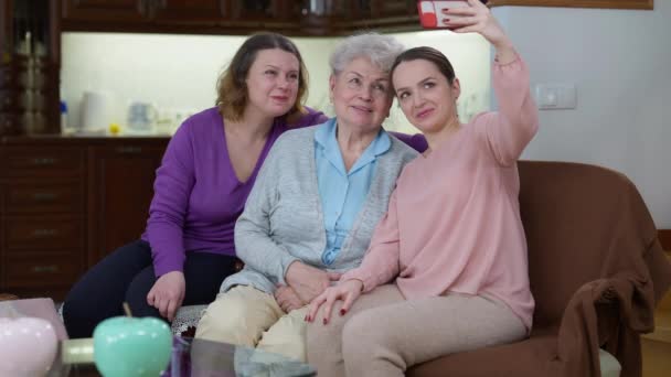 Lykkelig, vakker, ung kvinne som tar selfie med mor og bestemor hjem. Portrett av muntert smilende, kaukasisk datter som fotograferer minner på smarttelefonen. – stockvideo