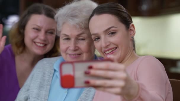 Portræt af munter glad kaukasiske unge kvinde tager selfie på smartphone med bedstemor og mor. Glædelig smilende smuk slank dame fotografere minder hjemme indendørs på ferie. – Stock-video